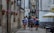 A group of hikers with backpacks and outdoor gear walks down a narrow cobblestone street between old stone buildings. Brightly colored flowers in pots are mounted on the wall, and there are several restaurants and cafes with outdoor seating areas featuring large umbrellas.
