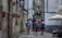 A group of hikers with backpacks and outdoor gear walks down a narrow cobblestone street between old stone buildings. Brightly colored flowers in pots are mounted on the wall, and there are several restaurants and cafes with outdoor seating areas featuring large umbrellas.