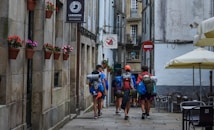 A group of hikers with backpacks and outdoor gear walks down a narrow cobblestone street between old stone buildings. Brightly colored flowers in pots are mounted on the wall, and there are several restaurants and cafes with outdoor seating areas featuring large umbrellas.