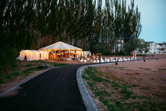 A large white tent is set up outdoors, surrounded by trees and open space. It seems to be part of a gathering or event, with people visible inside. The tent is illuminated with warm lights, creating a cozy atmosphere. A paved path leads up to the tent, and there are various decorations, possibly for an event or celebration. Nearby, there are some residential buildings visible in the background.
