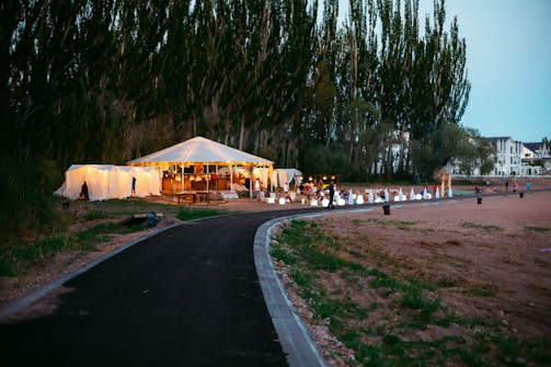 A large white tent is set up outdoors, surrounded by trees and open space. It seems to be part of a gathering or event, with people visible inside. The tent is illuminated with warm lights, creating a cozy atmosphere. A paved path leads up to the tent, and there are various decorations, possibly for an event or celebration. Nearby, there are some residential buildings visible in the background.