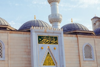 An ornate mosque exterior featuring large domes with dark grey tops and a central minaret with intricate detailing. The facade is adorned with decorative windows and Arabic calligraphy on green and gold panels, emphasizing architectural elegance.