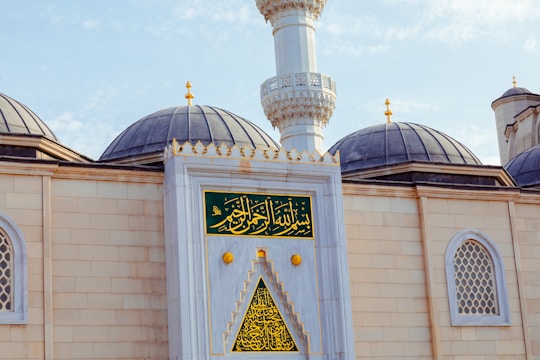 An ornate mosque exterior featuring large domes with dark grey tops and a central minaret with intricate detailing. The facade is adorned with decorative windows and Arabic calligraphy on green and gold panels, emphasizing architectural elegance.