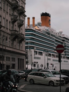 A large cruise ship passes by closely alongside an urban area with classic European architecture. Multiple cars and a motorcycle are visible in the foreground, navigating around a busy intersection as they drive past a 'no entry' sign. The ship's smokestack is emitting a small plume of smoke.