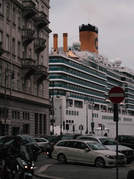 A large cruise ship passes by closely alongside an urban area with classic European architecture. Multiple cars and a motorcycle are visible in the foreground, navigating around a busy intersection as they drive past a 'no entry' sign. The ship's smokestack is emitting a small plume of smoke.