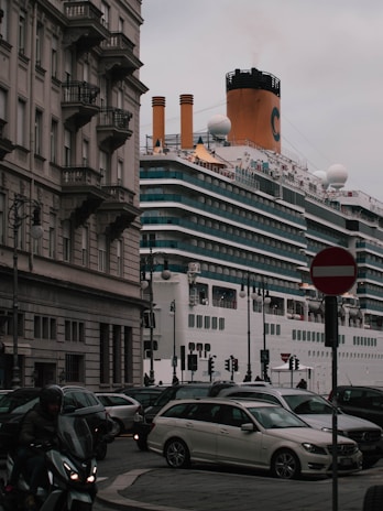 A large cruise ship passes by closely alongside an urban area with classic European architecture. Multiple cars and a motorcycle are visible in the foreground, navigating around a busy intersection as they drive past a 'no entry' sign. The ship's smokestack is emitting a small plume of smoke.
