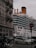 A large cruise ship passes by closely alongside an urban area with classic European architecture. Multiple cars and a motorcycle are visible in the foreground, navigating around a busy intersection as they drive past a 'no entry' sign. The ship's smokestack is emitting a small plume of smoke.
