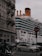 A large cruise ship passes by closely alongside an urban area with classic European architecture. Multiple cars and a motorcycle are visible in the foreground, navigating around a busy intersection as they drive past a 'no entry' sign. The ship's smokestack is emitting a small plume of smoke.
