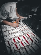 A person wearing a white chef's coat is meticulously decorating cookies shaped like gift boxes. The cookies are lined up on a baking tray, and the person is using a icing bag filled with red and orange icing. The gift box cookies have icing with bow designs, some of which are filled in with red color.