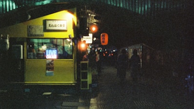 A dimly lit urban alley with a yellow structure prominently displayed. Neon signs reading 'LIVE ON AIR' and various Japanese characters are visible. People are walking in the shadows, adding a moody, mysterious atmosphere to the scene.
