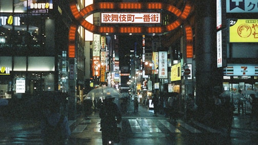 A neon-lit street in Tokyo at night, rain glistening on the pavement with blurred reflections.