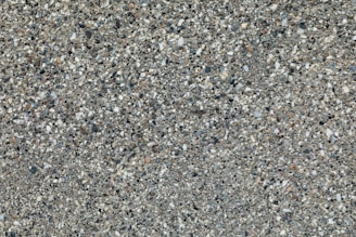 A textured surface composed of small pebbles and stones embedded in a rough, gray cement matrix. The stones vary in color, including shades of white, black, and brown, distributing randomly across the surface.