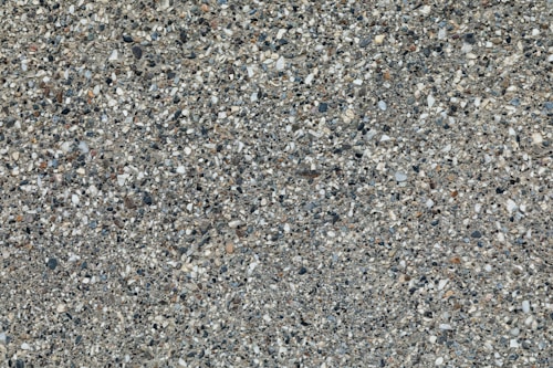 A textured surface composed of small pebbles and stones embedded in a rough, gray cement matrix. The stones vary in color, including shades of white, black, and brown, distributing randomly across the surface.