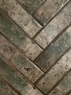 Close-up view of a herringbone pattern made from rectangular tiles that have a rough, textured surface. The tiles exhibit earthy tones with a mix of green and brown shades, giving an aged or rustic appearance.