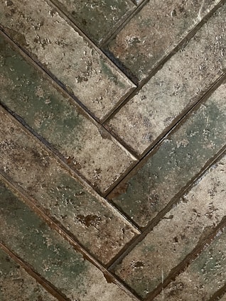 Close-up view of a herringbone pattern made from rectangular tiles that have a rough, textured surface. The tiles exhibit earthy tones with a mix of green and brown shades, giving an aged or rustic appearance.