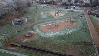 An aerial view of a playground in a park, featuring various play equipment such as swings, slides, and climbing structures. The ground is slightly dusted with frost, indicating a cold environment. Trees with frosted branches surround the area, and there is a path running through the park.