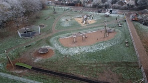 An aerial view of a playground in a park, featuring various play equipment such as swings, slides, and climbing structures. The ground is slightly dusted with frost, indicating a cold environment. Trees with frosted branches surround the area, and there is a path running through the park.