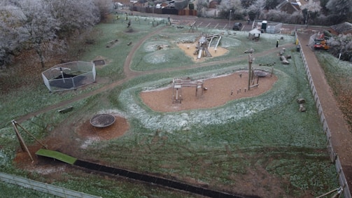 An aerial view of a playground in a park, featuring various play equipment such as swings, slides, and climbing structures. The ground is slightly dusted with frost, indicating a cold environment. Trees with frosted branches surround the area, and there is a path running through the park.