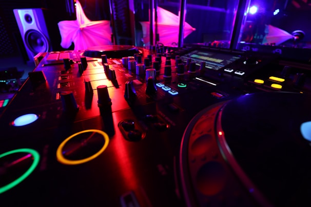 A close-up view of a DJ mixer and turntable setup in a dimly lit environment, featuring vibrant colored lights in the background. Various knobs, buttons, and displays are visible on the mixer, indicating a high-tech audio setup.