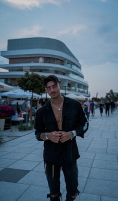 A person stands in front of a modern, multi-story building with a curved design. The scene is outdoors with a paved walkway and several people in the background. The individual is dressed in dark clothing with accessories, appearing casually stylish.