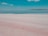 A vast landscape featuring a pink-hued flatland meeting a calm body of water that reflects the sky. A solitary person is walking across the expansive salt flat under a broad blue sky dotted with a few clouds.