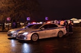 Two police cars are parked on a wet street at night with their lights on. Several people, including officers in reflective jackets, are standing near the vehicles. Street signs and trees are visible in the background.