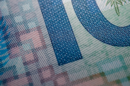 Close-up of a printed currency or security document featuring intricate patterns, detailed line work, and a large numeral. The design includes a color-shifting effect and a textured appearance, often seen in banknotes.