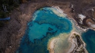 An aerial view of a natural wetland area featuring a bright blue body of water surrounded by dense, dry vegetation. The water appears to have intricate patterns formed by darker soil or algae. The surrounding landscape is characterized by brownish tones, indicating dry reeds or grasses.