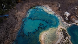An aerial view of a natural wetland area featuring a bright blue body of water surrounded by dense, dry vegetation. The water appears to have intricate patterns formed by darker soil or algae. The surrounding landscape is characterized by brownish tones, indicating dry reeds or grasses.