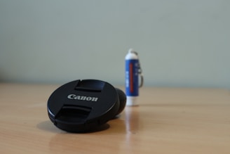 A black Canon lens cap is positioned on a wooden surface in the foreground, with a tube of glue or lip balm standing upright in the background.