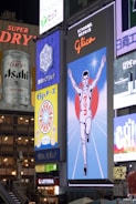 A bustling urban scene featuring large, illuminated billboards and advertisements. Prominent among them is a vintage-style sign depicting a running athlete with a red circle behind him, known as the Glico running man. Other visible ads include those for beverages and snacks, all contributing to a vibrant, colorful mix of visuals.