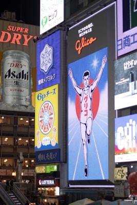A bustling urban scene featuring large, illuminated billboards and advertisements. Prominent among them is a vintage-style sign depicting a running athlete with a red circle behind him, known as the Glico running man. Other visible ads include those for beverages and snacks, all contributing to a vibrant, colorful mix of visuals.
