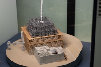 A detailed architectural model of a building under construction with scaffolding and structural framework visible. The model includes a rectangular base and various sections made of wood and metal. The setting is indoors, with the model displayed on a round platform.