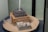 A detailed architectural model of a building under construction with scaffolding and structural framework visible. The model includes a rectangular base and various sections made of wood and metal. The setting is indoors, with the model displayed on a round platform.