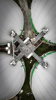 An aerial view of four large circular industrial tanks connected by metal walkways, forming a cross shape. The tanks are covered with a light gray material and bordered by green accents. The surrounding area is composed of gravel and grass, creating a contrast with the industrial structures.