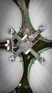 An aerial view of four large circular industrial tanks connected by metal walkways, forming a cross shape. The tanks are covered with a light gray material and bordered by green accents. The surrounding area is composed of gravel and grass, creating a contrast with the industrial structures.