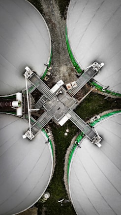 An aerial view of four large circular industrial tanks connected by metal walkways, forming a cross shape. The tanks are covered with a light gray material and bordered by green accents. The surrounding area is composed of gravel and grass, creating a contrast with the industrial structures.