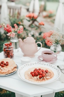 A beautifully arranged outdoor brunch setup on a white table with pancakes topped with fresh raspberries. A pink teapot and a jar filled with raspberries are placed alongside a cup of tea. Flower arrangements with pink and white blossoms enhance the serene setting.
