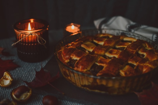 A rustic setting with a freshly baked pie featuring a golden brown lattice crust inside a glass dish. Two lit candles in dark, textured holders create a warm, cozy atmosphere. The scene is complemented by scattered autumn leaves and chestnuts on a woven fabric background.