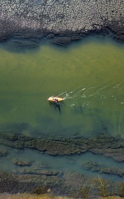Aerial view of a small yellow kayak being paddled by two people on a river with clear, greenish water. The surrounding riverbed reveals rocky and textured terrain, creating an interesting contrast between the water and land.