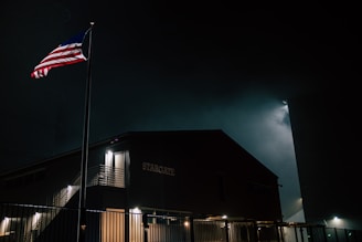 A building labeled 'STARGATE' is illuminated softly in the night, with an American flag waving on a pole nearby. The scene is enveloped in darkness, with a faint glow behind the structure, potentially from ambient city lights or moonlight. A metal fence encloses the area.