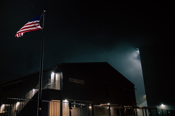 A building labeled 'STARGATE' is illuminated softly in the night, with an American flag waving on a pole nearby. The scene is enveloped in darkness, with a faint glow behind the structure, potentially from ambient city lights or moonlight. A metal fence encloses the area.