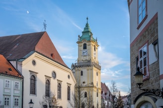 A historic town square featuring a prominent clock tower with a green patina roof. The sky is clear with visible daylight, and a few bare trees line the street. Surrounding buildings have classic architectural details with various roof shapes and a mix of earth tones and pastel colors.