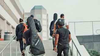 A group of people is walking up a set of stairs in an urban area. Some individuals are carrying musical instrument cases on their backs. The background includes modern, multi-story buildings and railings, with a clear sky.