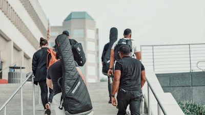A group of people is walking up a set of stairs in an urban area. Some individuals are carrying musical instrument cases on their backs. The background includes modern, multi-story buildings and railings, with a clear sky.
