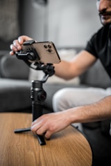 A person is adjusting a smartphone mounted on a stabilizer or gimbal, which is placed on a wooden tabletop. The focus is on the hand and the device, capturing the detail of the phone and stabilizer. The background is softly blurred, giving a sense of a cozy, indoor setting.