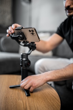 A person is adjusting a smartphone mounted on a stabilizer or gimbal, which is placed on a wooden tabletop. The focus is on the hand and the device, capturing the detail of the phone and stabilizer. The background is softly blurred, giving a sense of a cozy, indoor setting.