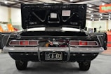 A classic black car with an open trunk is displayed indoors. The car has a license plate from Georgia, marked 15-A-473, 1969 Peach State. The vehicle features dual exhausts and a distinctive rear tail light design. The exhibition hall is well-lit with visible signage and other vehicles in the background.