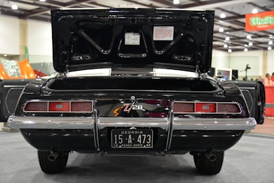 A classic black car with an open trunk is displayed indoors. The car has a license plate from Georgia, marked 15-A-473, 1969 Peach State. The vehicle features dual exhausts and a distinctive rear tail light design. The exhibition hall is well-lit with visible signage and other vehicles in the background.