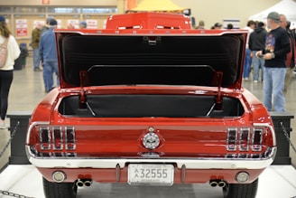 A classic red car with its trunk open is displayed indoors. Several people are visible in the background, suggesting a car show or exhibition setting. The car's license plate indicates it is an antique vehicle from Tennessee. The setting appears busy with various attendees walking around the venue.